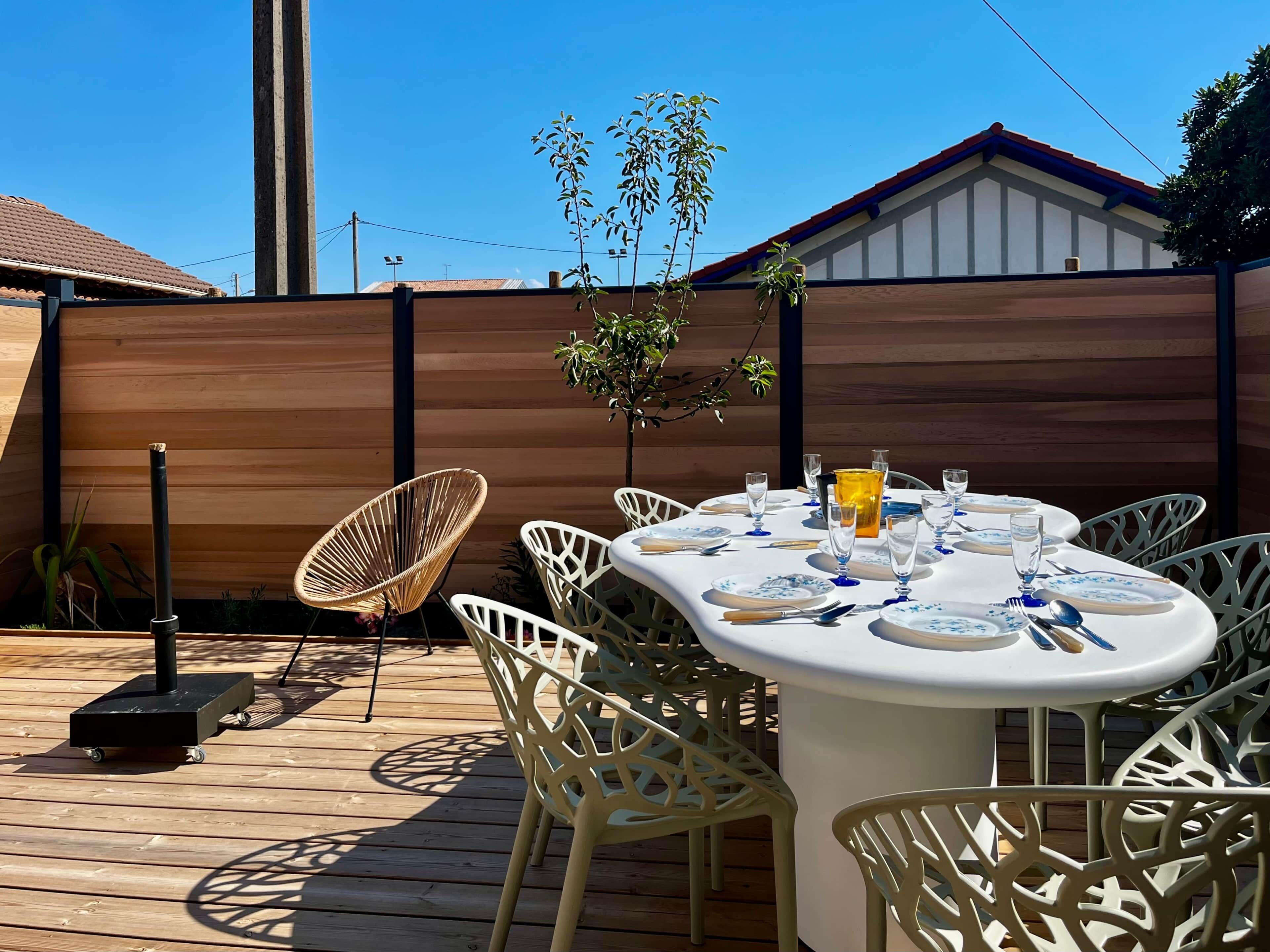 An outdoor dining area with a table set for four, surrounded by white chairs, on a wooden deck.