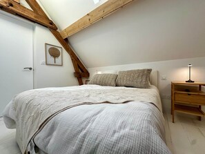 Ein Schlafzimmer mit Schrägwand, einem Bett mit strukturierter Decke, zwei Kissen, einer Nachttischlampe und einem Holznachttisch.