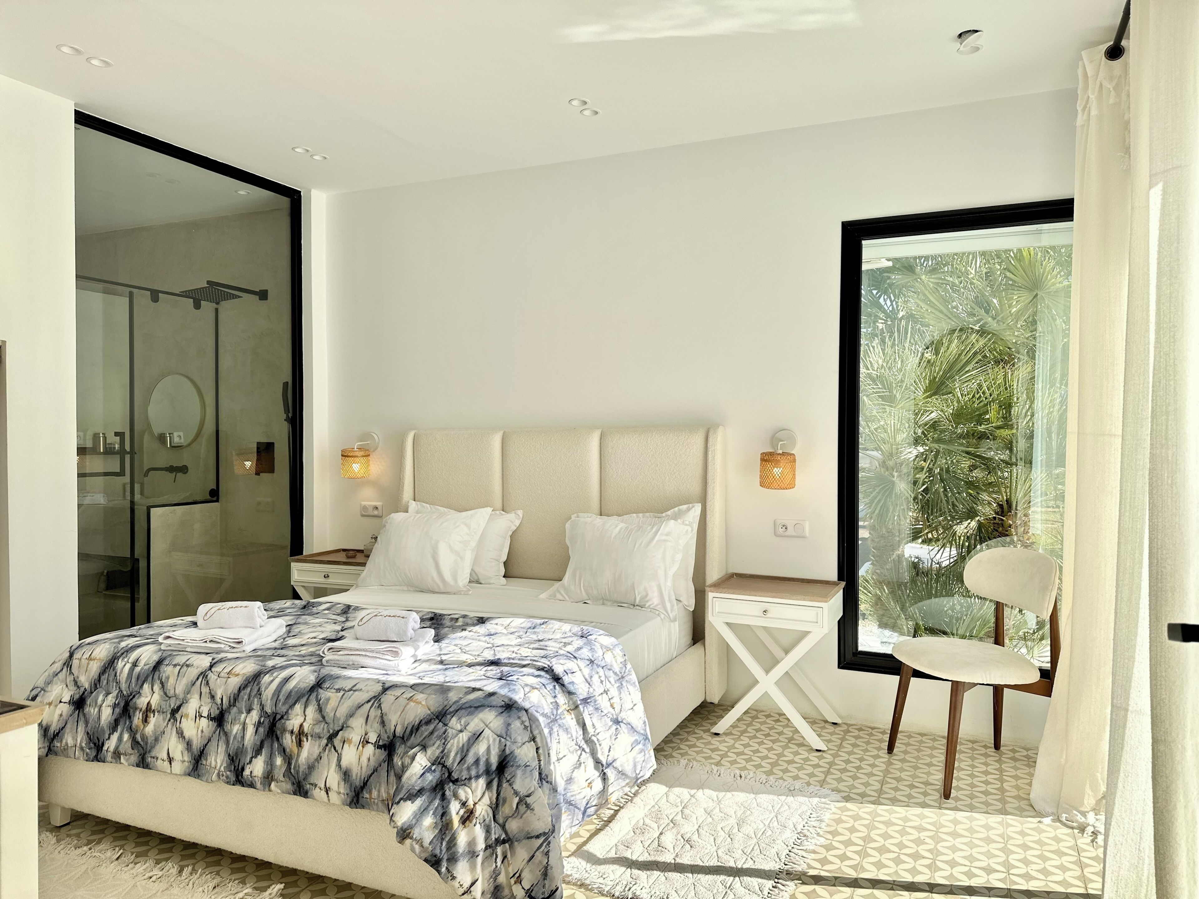 A modern bedroom with a large bed, a glass shower enclosure, and a view of greenery through a sliding door.