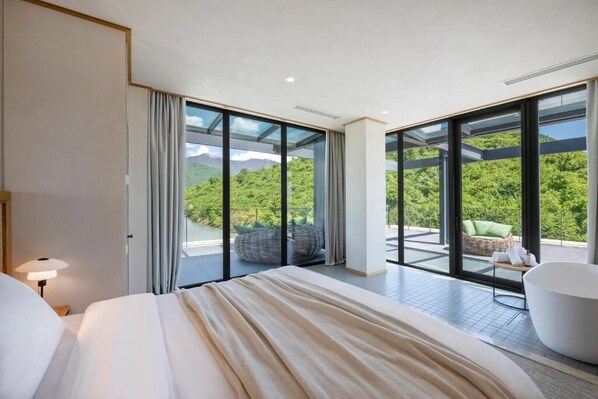 A modern hotel room with a large bed, a view of lush greenery, and a balcony with outdoor seating.