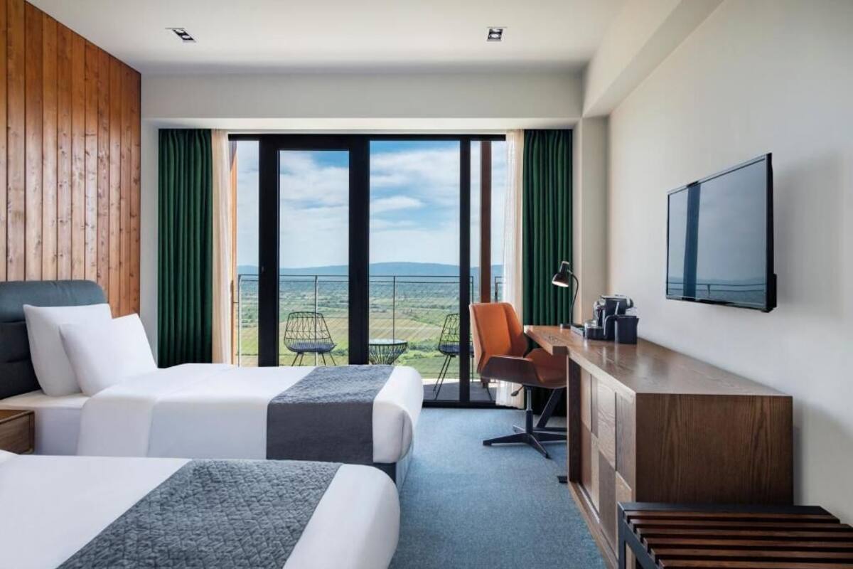 A hotel room with two beds, a desk, a chair, a TV, and a view of the outdoors.