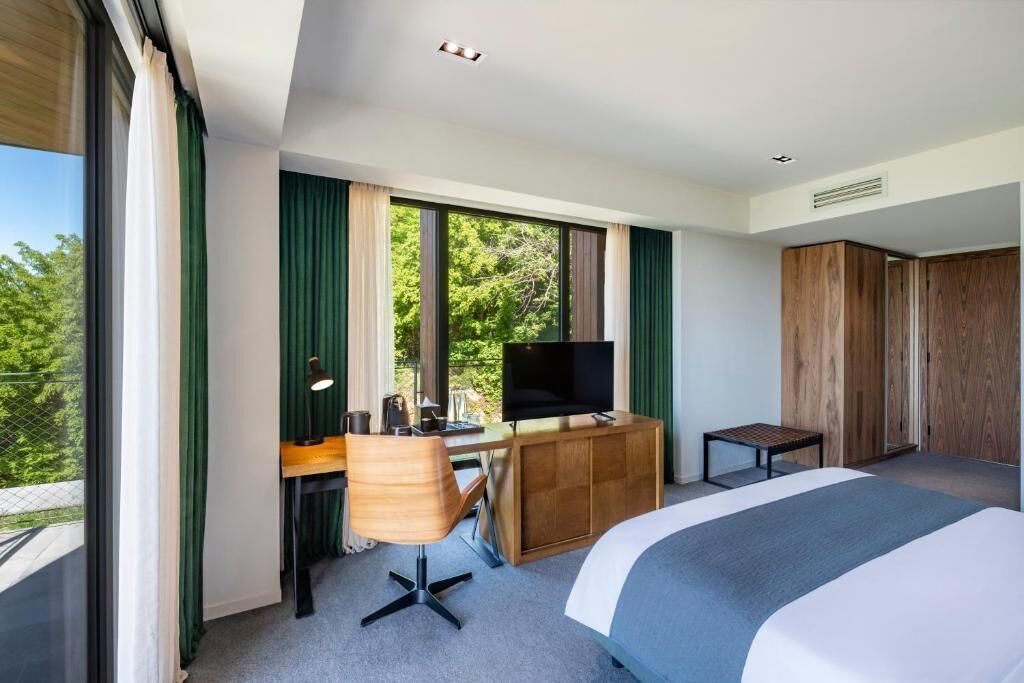 A modern hotel room with a large bed, a desk with a TV, and a view of greenery outside.