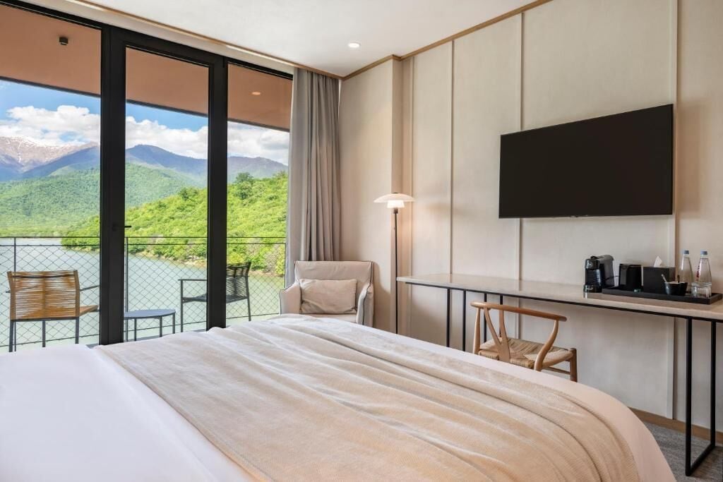 A hotel room with a large bed, a desk, a chair, a TV, and a balcony with a mountain view.