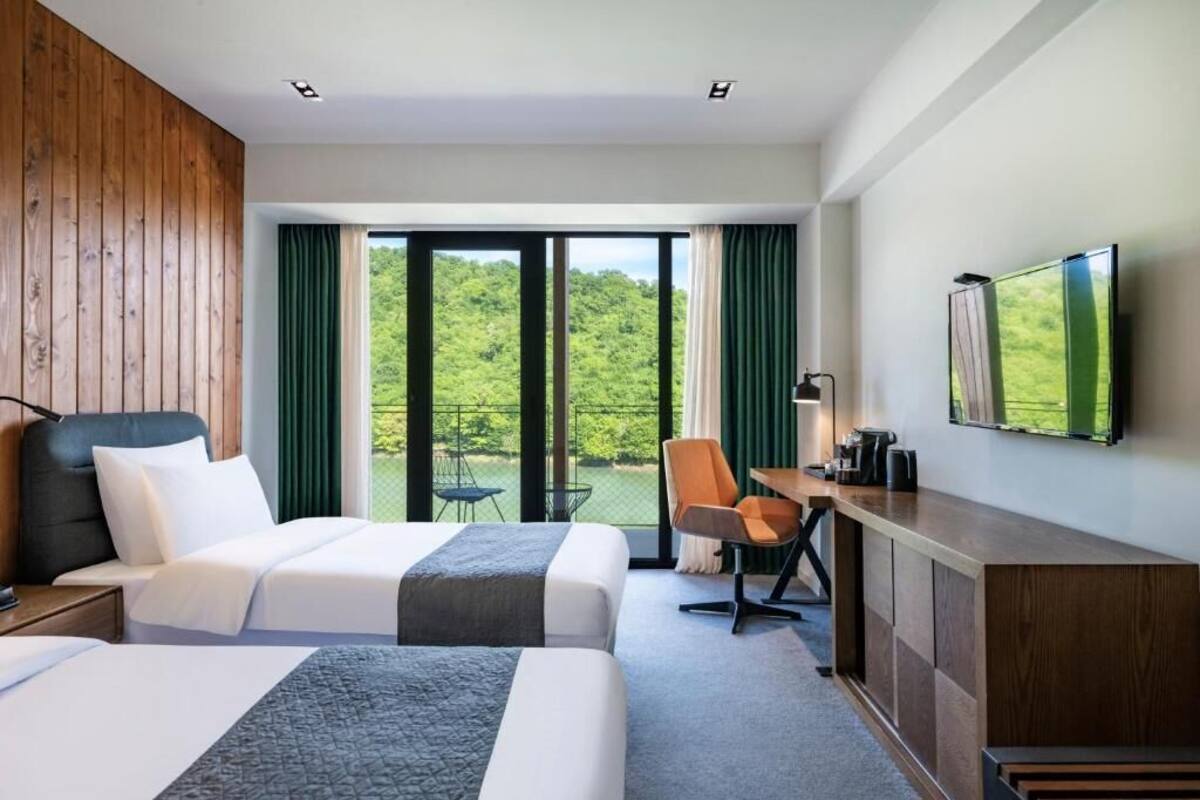 A hotel room with two beds, a desk, a chair, a TV, and a balcony with a view of greenery.