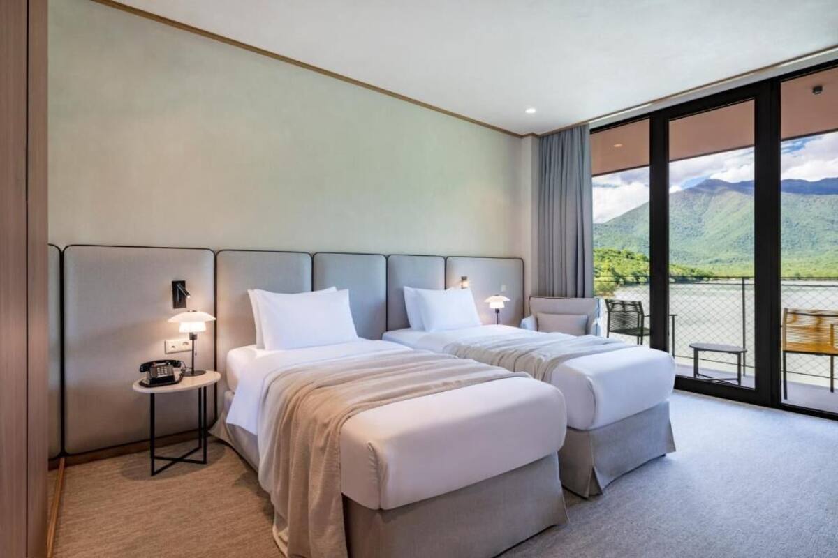 A hotel room with two beds, a small table with a lamp, and a view of mountains through large windows.