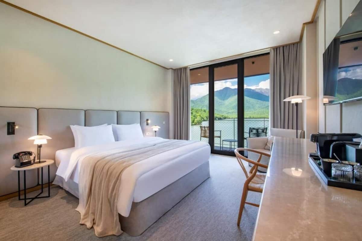 A hotel room with a large bed, a desk with a chair, a phone, and a view of mountains through the window.