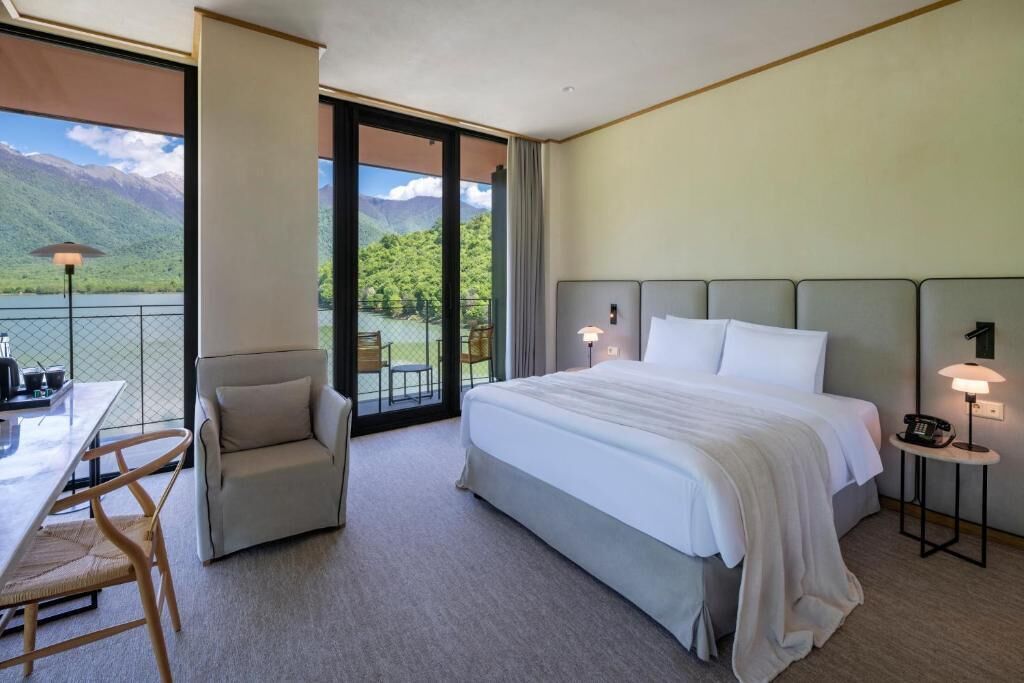 Habitación de hotel con una cama grande, un escritorio con silla, una mesita de noche con lámpara y vistas a montañas a través de un ventanal.