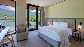 Habitación de hotel con una cama grande, un escritorio con silla, una mesita de noche con lámpara y vistas a montañas a través de un ventanal.