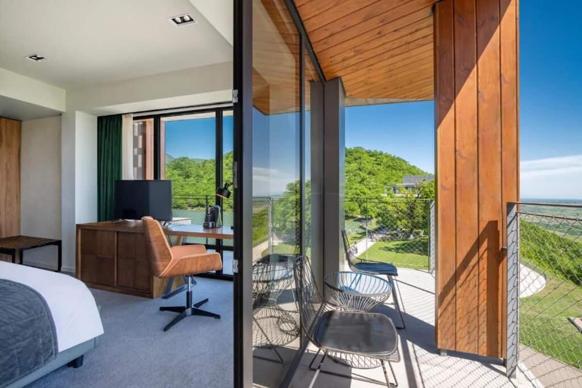 A modern hotel room with a large bed, a desk with a chair, a TV, and a balcony with a view of greenery.
