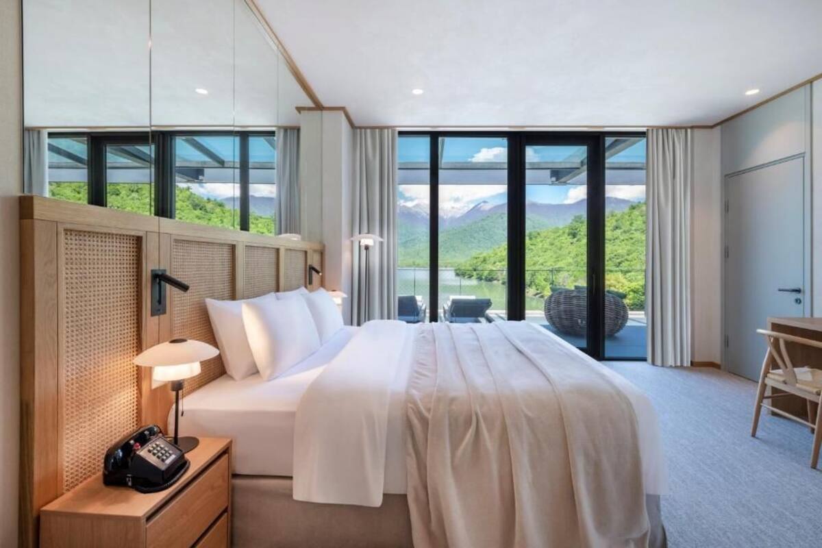 A modern hotel room with a large bed, a desk, and a view of mountains through floor-to-ceiling windows.