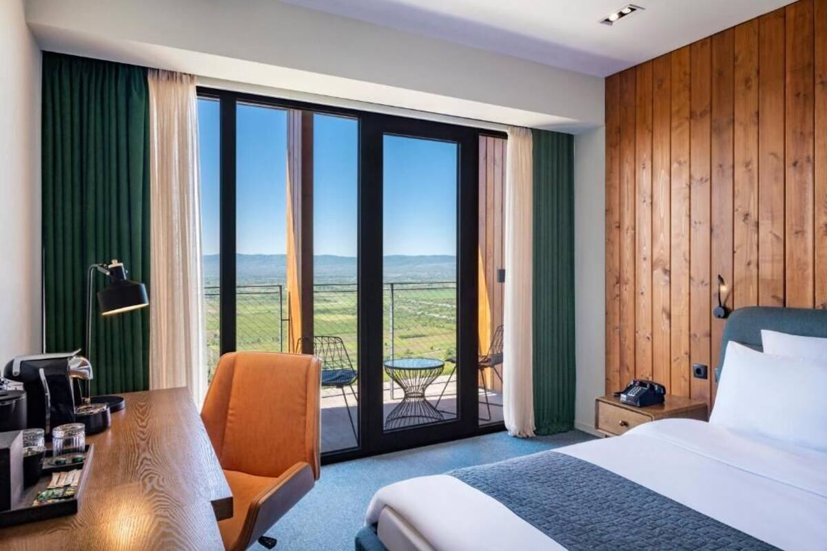 A hotel room with a large bed, a desk with a lamp, a chair, and a view of a green landscape through a sliding glass door.