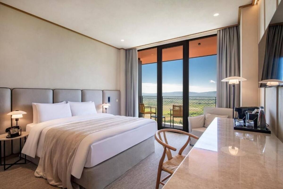 A hotel room with a large bed, a desk, a chair, and a view of the landscape through the window.