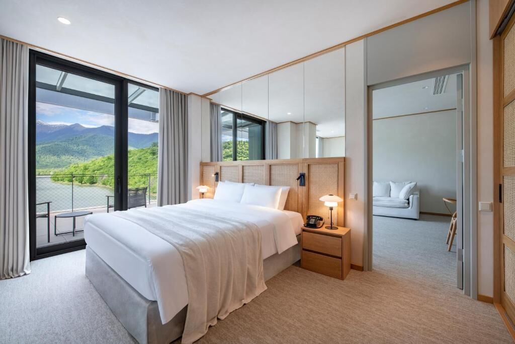 A modern hotel room with a large bed, a bedside table with a lamp, a view of mountains through large windows, and a balcony.