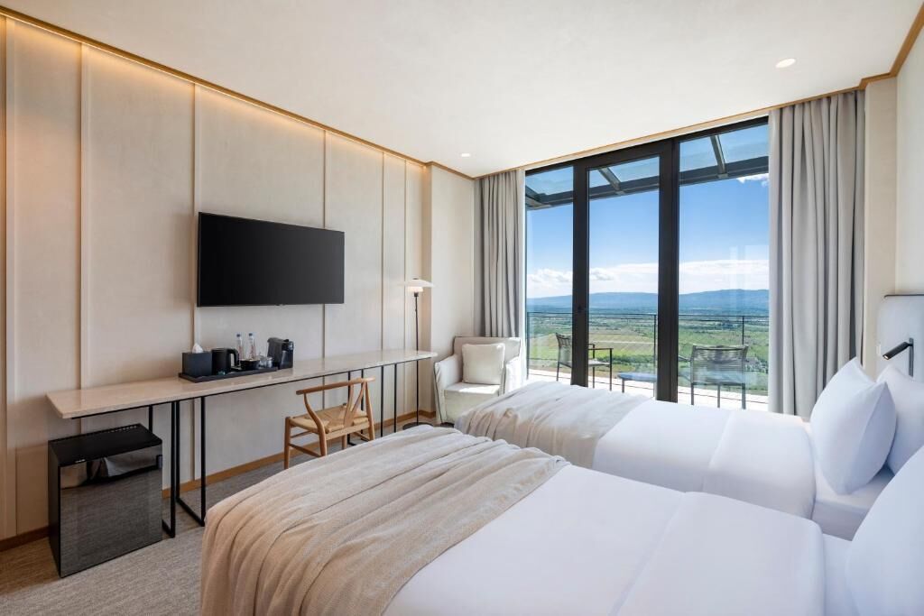 A hotel room with two beds, a desk, a TV, and a large window with a view of the landscape.