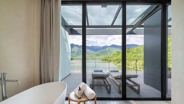 A modern bathroom with a freestanding bathtub, a wooden stool with towels, and a large sliding glass door leading to a balcony with outdoor seating and a mountain view. - Kvareli Lake Resort (Kvareli)