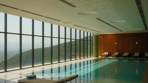 A modern indoor swimming pool with clear glass walls, lounge chairs, and a view of a mountainous landscape. - Kvareli Lake Resort (Kvareli)
