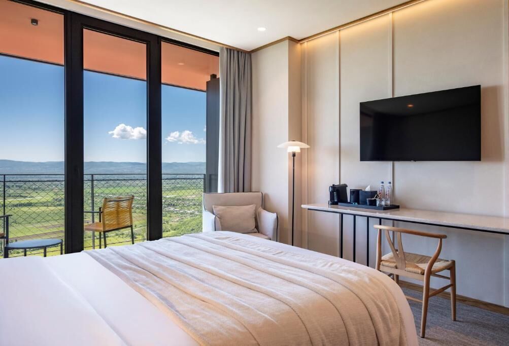 Ett hotellrum med en säng, ett skrivbord, en stol, en TV och en balkong med utsikt över landskapet.