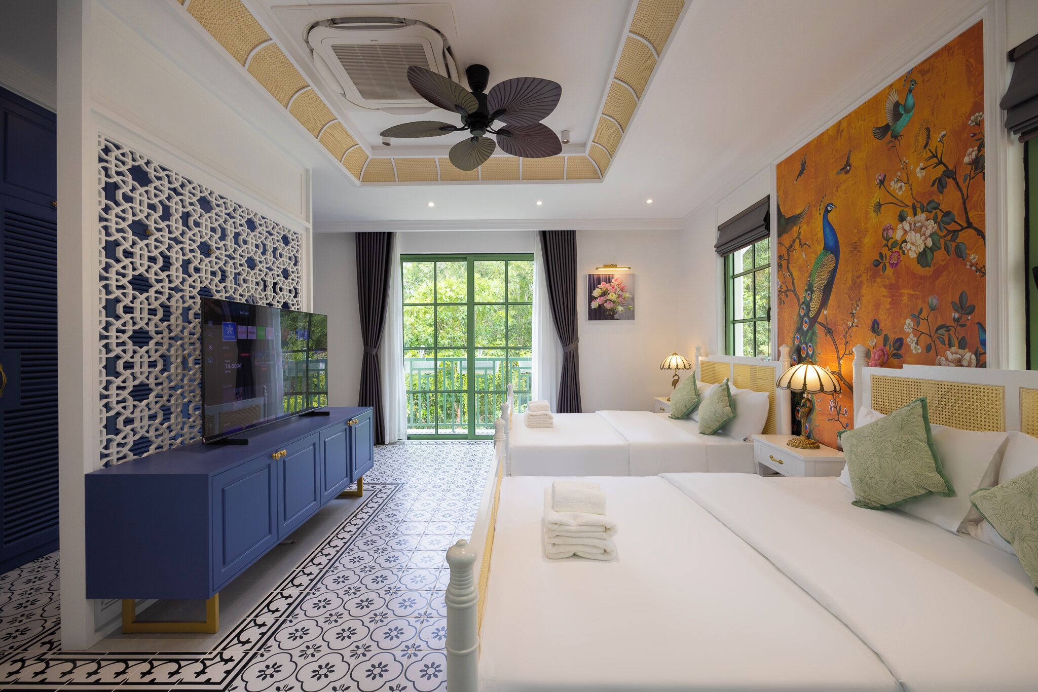 Una camera d'albergo con un letto grande, una TV su un mobile azzurro, una ventola a soffitto, una finestra con tende e un affresco al muro.