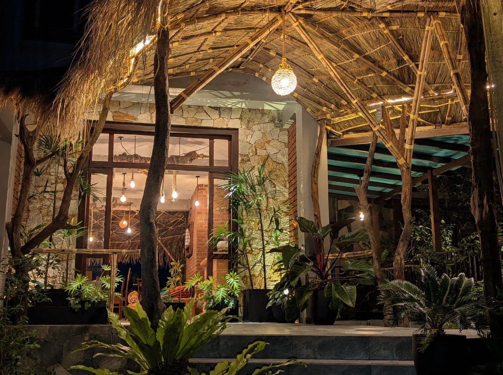 Una estructura con techo de paja, pared de piedra y ventanas amplias, rodeada de plantas tropicales.