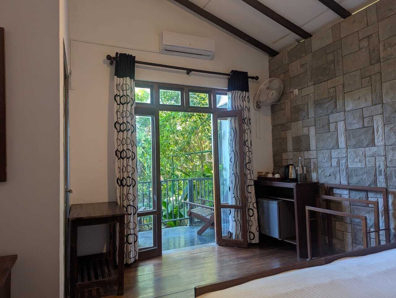 A room with a stone wall, a wooden table, and a view of a balcony with greenery.