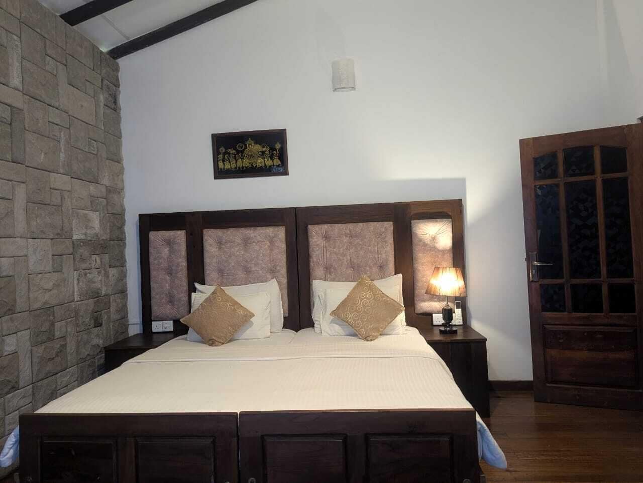 Quarto com uma cama grande, duas mesas de cabeceira, parede de pedra, porta de madeira e um quadro na parede.