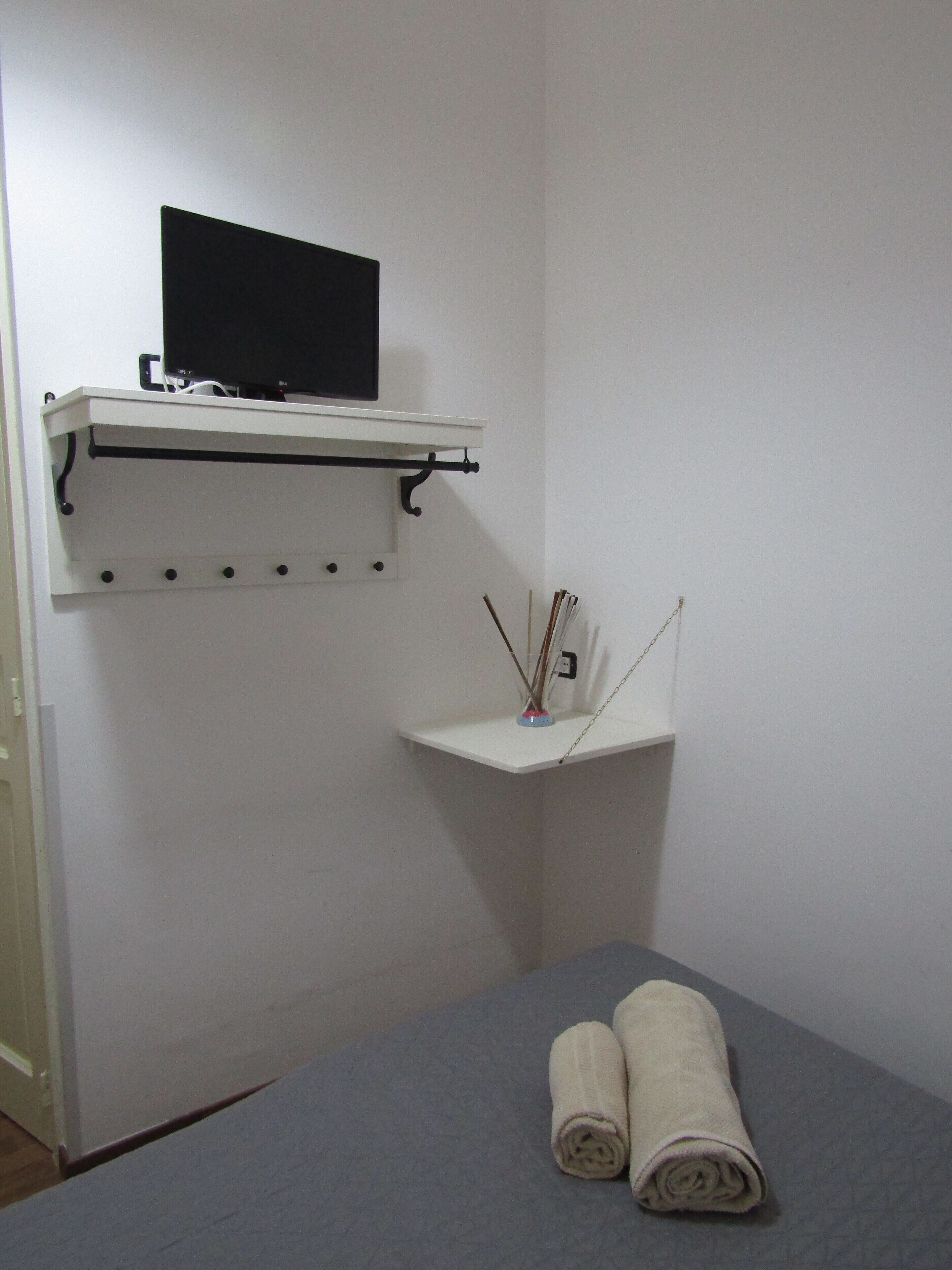 A room with a bed, a mounted TV, a small shelf with a vase, and a towel on the bed.