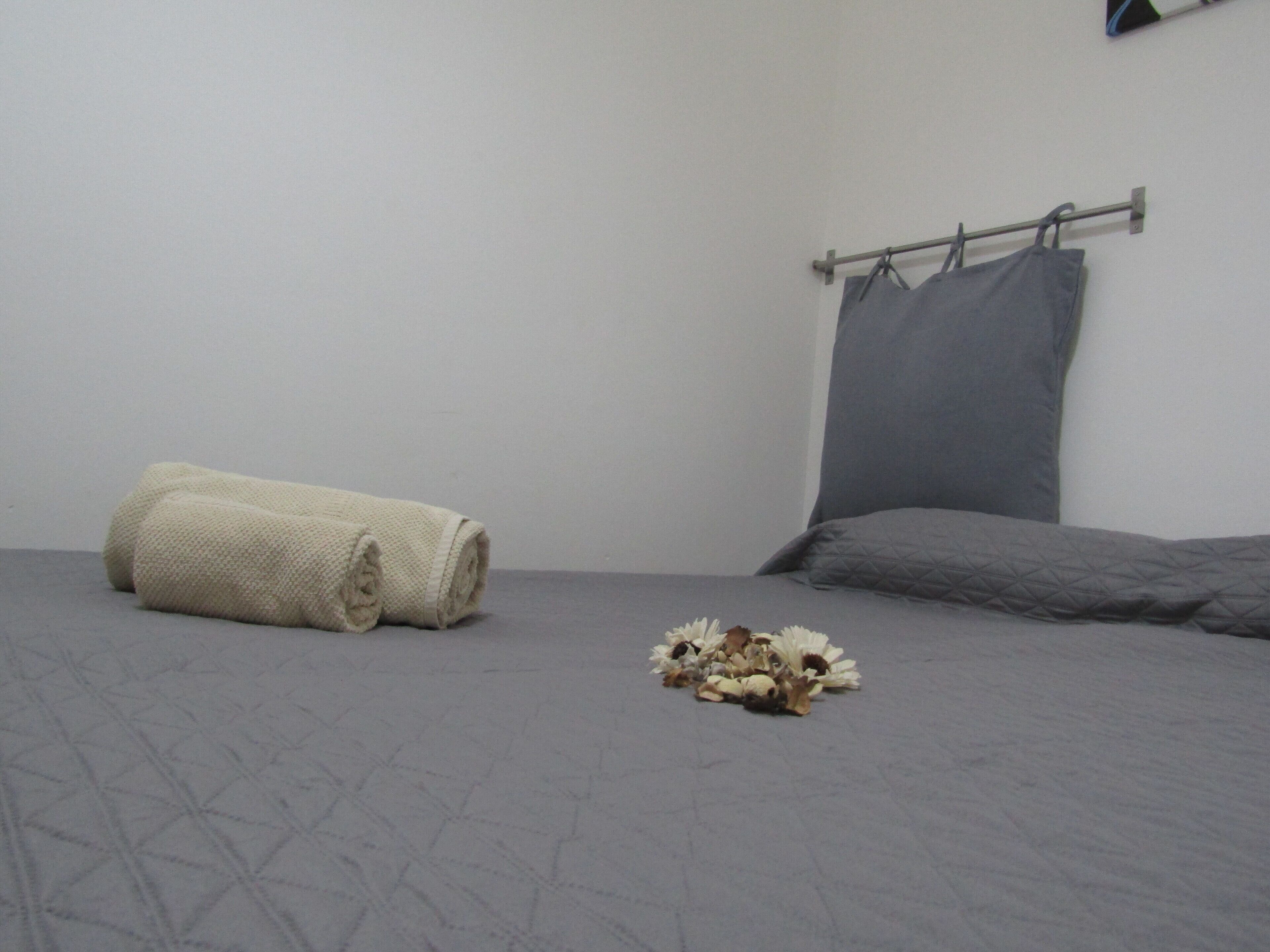 Une chambre avec un lit gris, un oreiller gris et une couverture beige enroulée sur le sol.