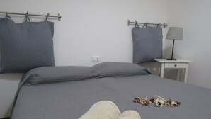 A neatly made bed with grey bedding and pillows, a small bedside table with a lamp, and a power outlet on the wall. - The White Room Courtyard (Scicli)