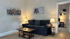 A living room with a dark-colored sofa, two side tables with lamps, a coffee table, and two wall-mounted paintings. - Elm Corner, 2 bedroom family house in Historic Loveland (Loveland)