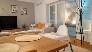 A modern living room with a wooden dining table, white chairs, a sofa with colorful cushions, a television, and a large window with sheer curtains. - Joy City Stay Madgearu 2 (Timișoara)