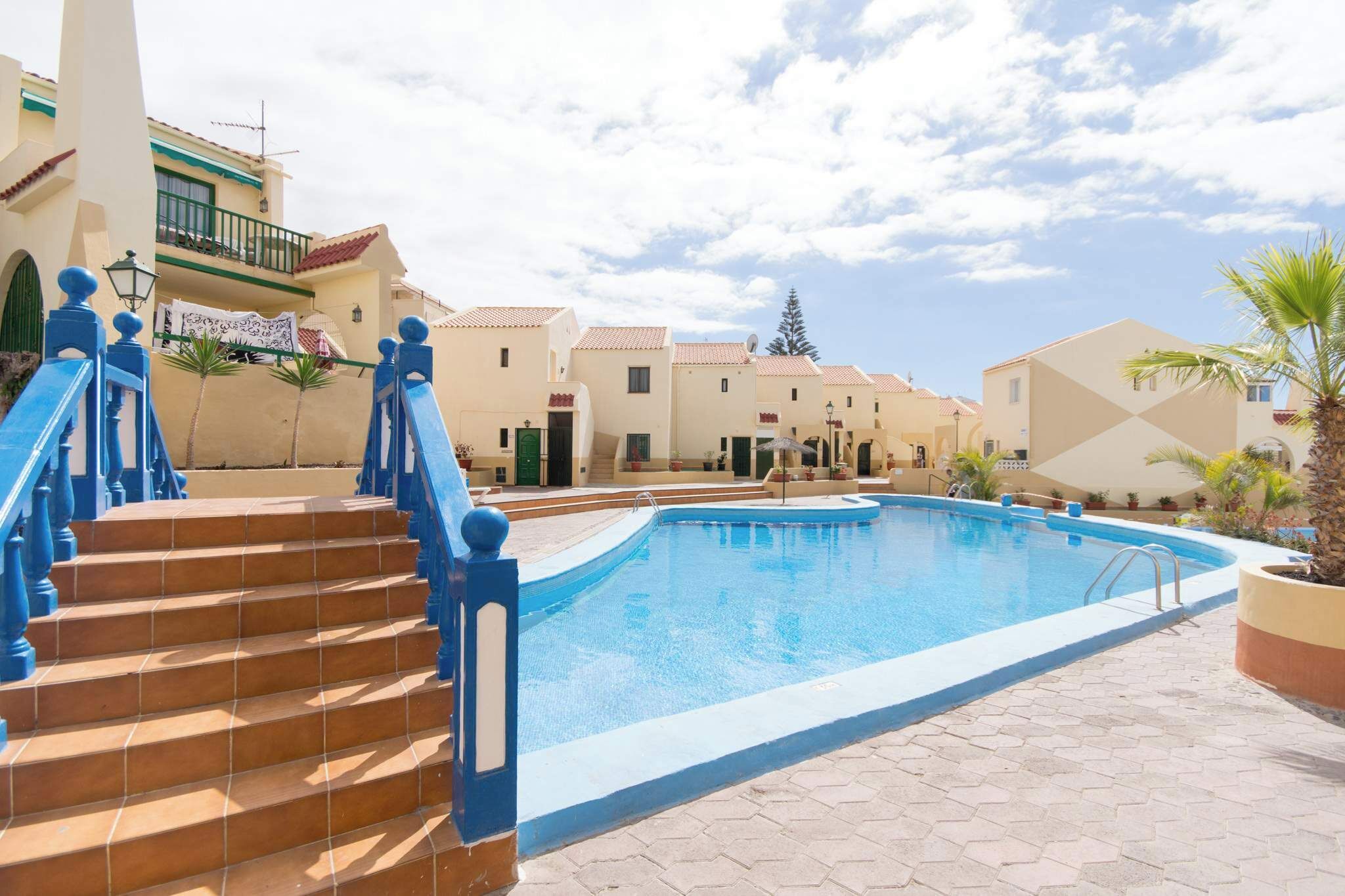 A swimming pool with blue-tiled steps leading up to it, surrounded by buildings with balconies and a clear sky.