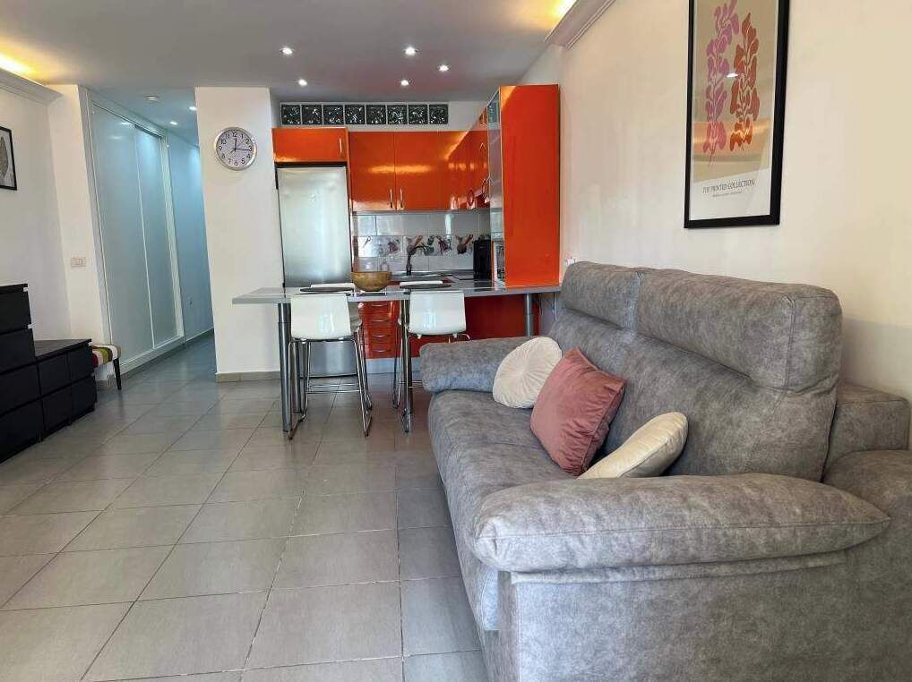 A modern living room with a grey sofa, a dining table with chairs, and a kitchen area with orange cabinets.