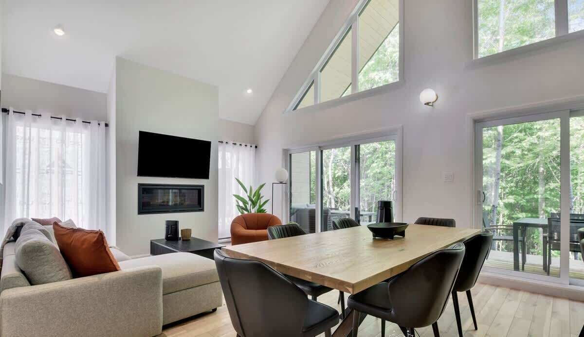A modern living room with a sofa, a dining table with chairs, a TV mounted on the wall, and large windows.