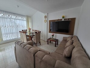 Una sala de estar con un sofá, una mesa de comedor con sillas, un televisor montado en la pared y una ventana con cortinas.