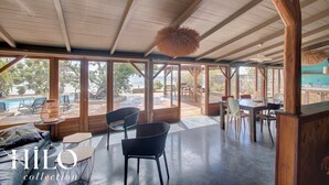 Ruime, overdekte ruimte met een hoge plafond, grote ramen en uitzicht op de omgeving. Er staan meerdere stoelen en tafels klaar voor eten of gezellig samenzijn.