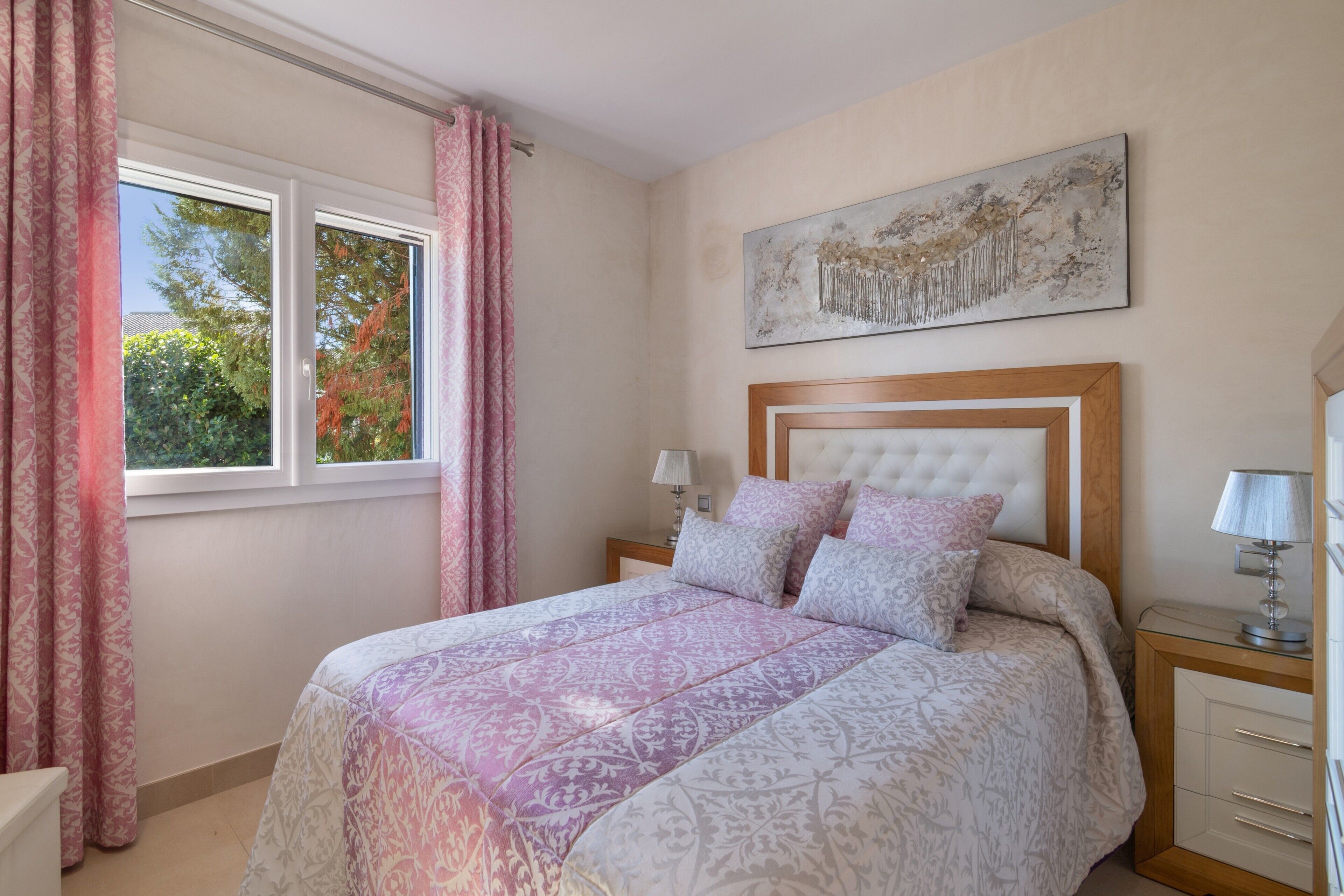 A bedroom with a bed, a nightstand, a lamp, a window with curtains, and a wall-mounted artwork.