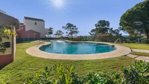 A swimming pool surrounded by a well-maintained lawn, with a clear sky and a building in the background. - Islantilla Golf Holiday Home with Private Terrace, Wi-Fi, and Air Conditioning (Huelva)