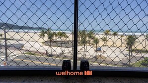 A view through a chain-link fence of a beach with palm trees, a road, and a building in the distance. - Apartamento na Praia Grande com vista mar (Praia Grande)