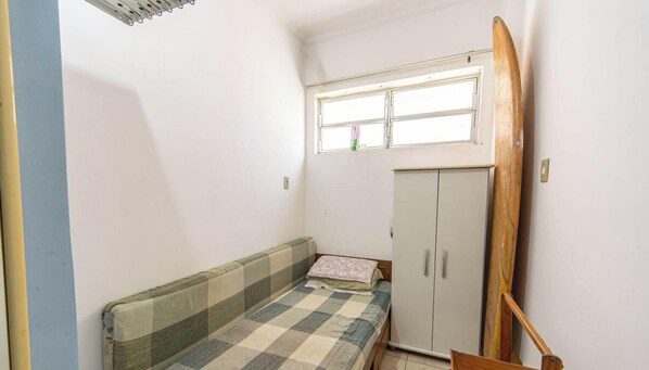 Una stanza piccola e arredata in modo essenziale, con un letto singolo, un armadio e una tavola da surf appoggiata al muro.