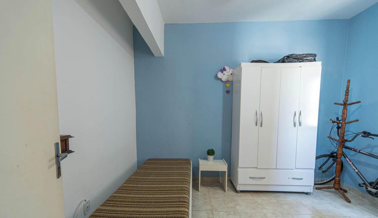 Une petite pièce avec un lit, une armoire blanche, une petite table avec une plante et un vélo.