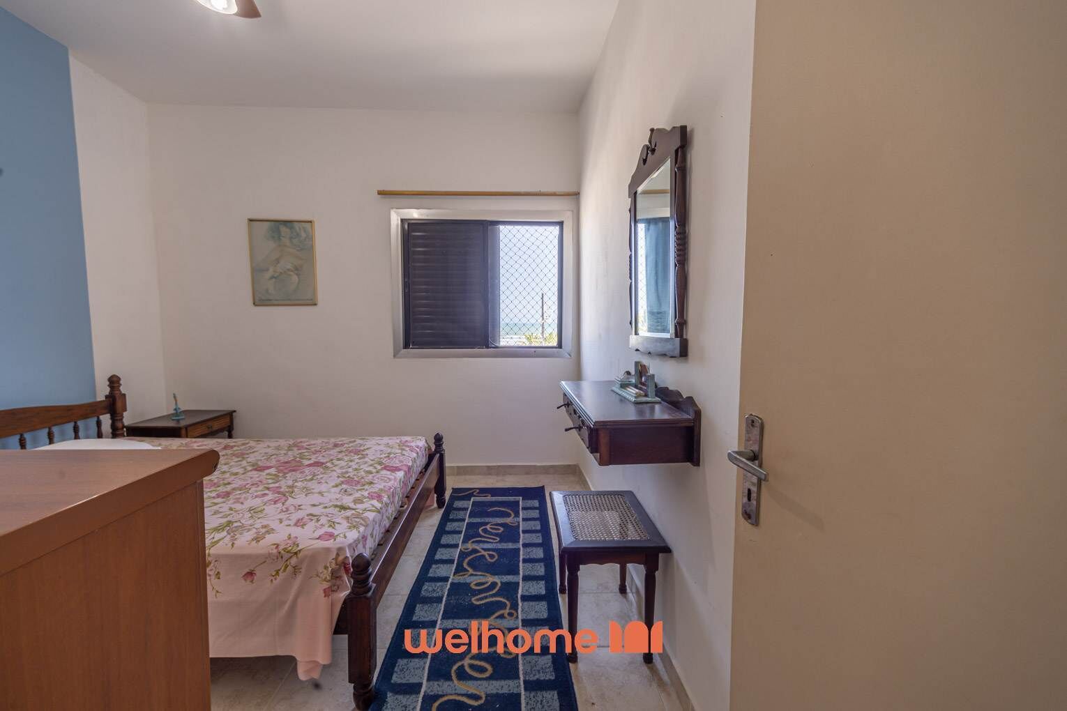 Une chambre à coucher comprenant un lit, une petite table, une table de chevet, une fenêtre avec des stores, un miroir et un paillasson.