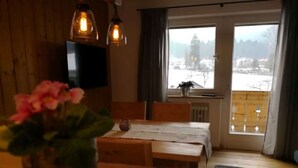 A cozy room with a wooden table, a vase with flowers, a clock, and a view of a snowy landscape through a large window. - Apartment in Grainau am Badersee (Grainau)