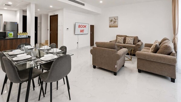 Ein modernes Hotelzimmer mit Essbereich, Sofas, einem Fernseher und einem Schild mit der Aufschrift 'BOOK ME'.
