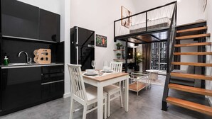 Una cocina moderna con gabinetes negros, un comedor con una mesa de madera y sillas blancas, un refrigerador negro y una escalera que sube a un nivel superior.
