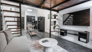 Una sala moderna con un sofá, una mesa redonda de centro, un televisor montado en la pared y una cocina al fondo.