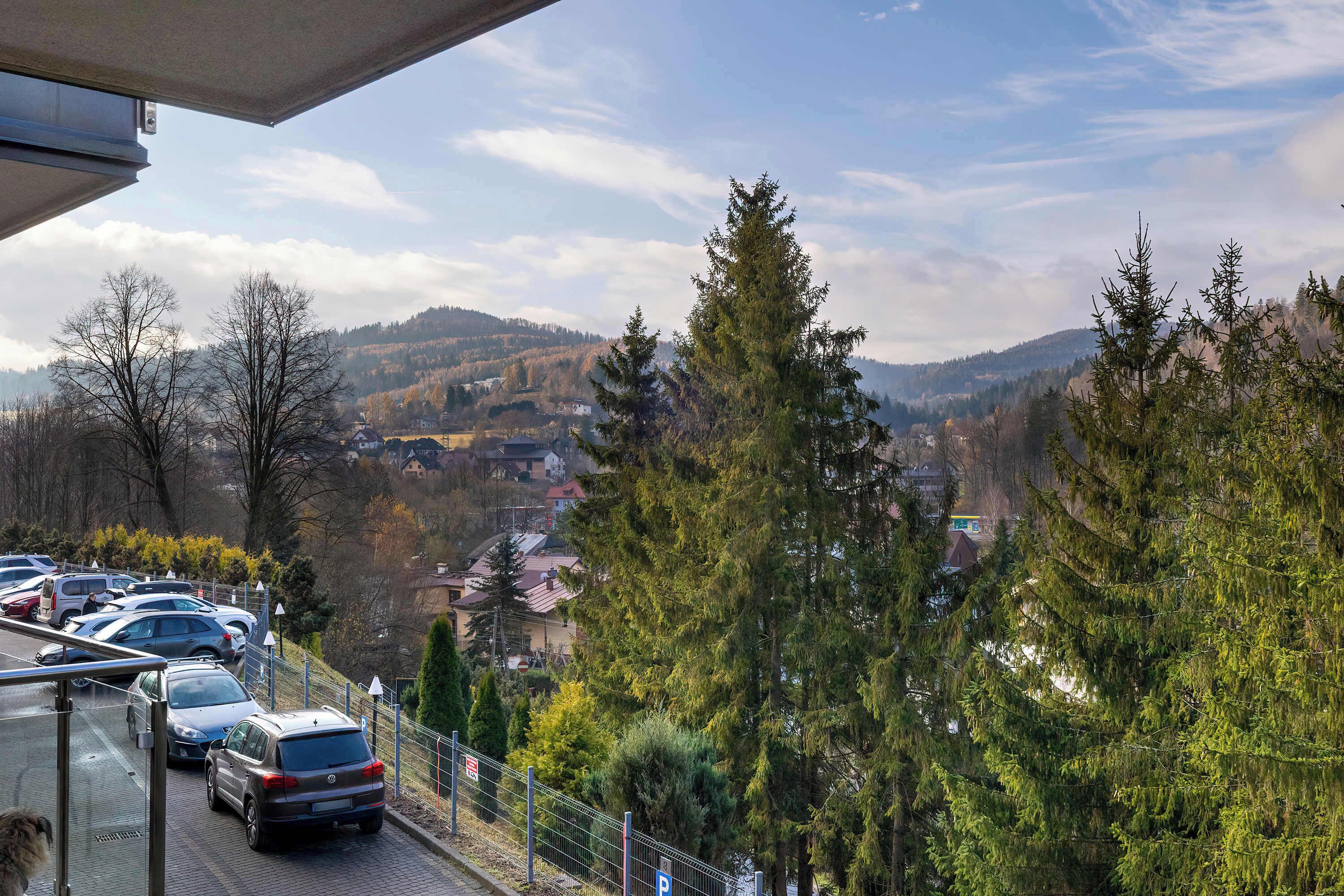 Un espace de stationnement avec des voitures, une clôture et une vue sur un paysage vallonné, avec des arbres et des bâtiments.