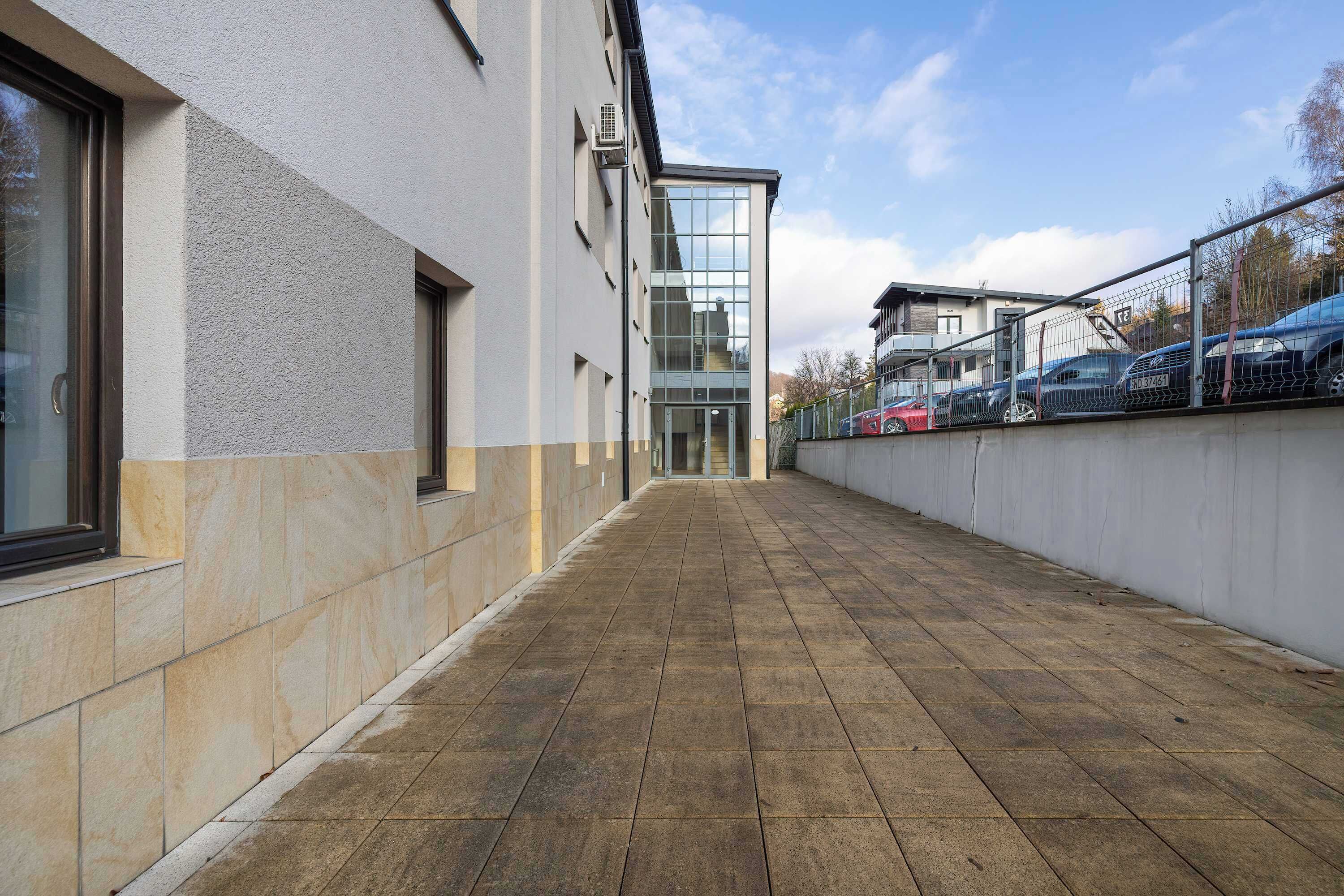 A corridor with a tiled floor, a building with large windows, and parked cars along the side.