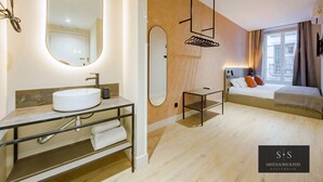 Ein modernes Hotelzimmer mit einem großen runden Spiegel, einem runden Waschbecken, einem kleinen Nachttisch, einem Bett mit Kissen und Blick auf die Stadt durch das Fenster.