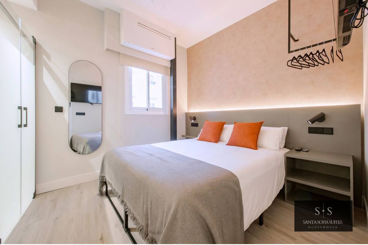 A modern bedroom with a large bed, a bedside table, a mirror, and a wall-mounted light fixture.