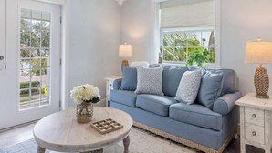 Un salon avec un canapé bleu, deux tables d’appoint avec des lampes, une table basse ronde ornée d’un vase et d’un plateau, et une grande fenêtre avec des stores.